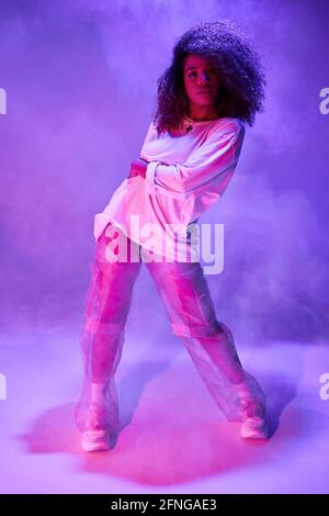 Tutta la lunghezza si adatta giovane ballerina afroamericana in sciolto indossare un abbigliamento informale guardando la macchina fotografica mentre si balla in uno studio buio nelle luci al neon Foto Stock