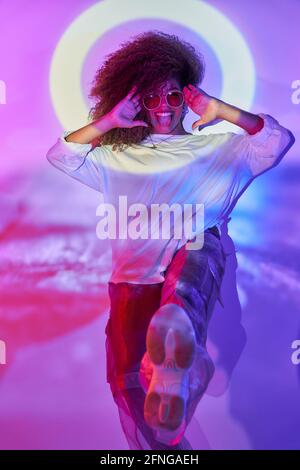 Giovane e sicura ballerina afro-americana in stile informale e gli occhiali da sole che ballano puntando i piedi verso la macchina fotografica che fa i volti in lig neon Foto Stock