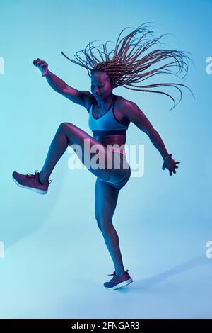 Forte atleta afroamericana in abbigliamento sportivo con il volo Afro trecce e bicipiti che guardano in su nella luce del neon Foto Stock