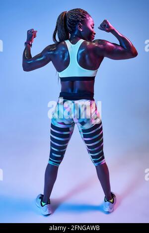 Vista posteriore di anonimo muscoloso afro-americano atleta femminile con corpo sudato che mostra bicipiti su sfondo blu Foto Stock