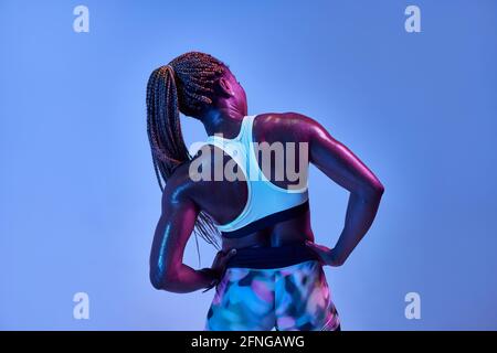 Vista posteriore di anonimo muscoloso afro-americano atleta femminile con corpo sudato che mostra bicipiti su sfondo blu Foto Stock