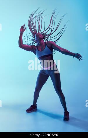 Forte atleta afroamericana in abbigliamento sportivo con il volo Afro trecce e bicipiti che guardano in su nella luce del neon Foto Stock