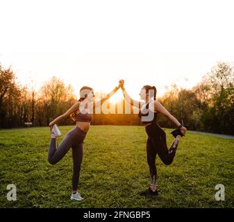 Vista laterale di donne atletiche sorridenti che tengono le mani e. allungare le gambe mentre si sostengono l'una con l'altra durante l'allenamento nel parco al tramonto Foto Stock