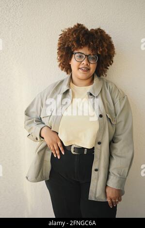 Bella felice sorridente curvy Plus taglia African nero donna afro capelli in posa in T-shirt beige, jeans, occhiali eleganti su sfondo beige. Imperfezione del corpo, concetto di diversità positiva del corpo di accettazione Foto Stock