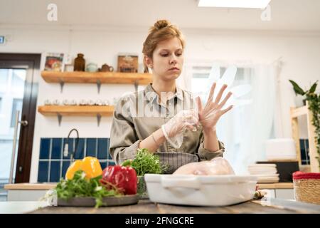 Donna che indossa guanti trasparenti e guarda verso il basso contro freschi peperoni con erbe e pollo crudo in teglia a. casa Foto Stock