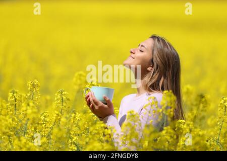 Profilo di una donna felice rilassante respirare aria profondamente fresca e tenendo una tazza di caffè in un campo giallo Foto Stock