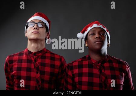 Due amici da uomo che indossano cappelli di Natale Babbo Natale e abbinamenti camicie rosse in piedi fianco a fianco guardando verso l'alto con espressioni gravi contro una g Foto Stock
