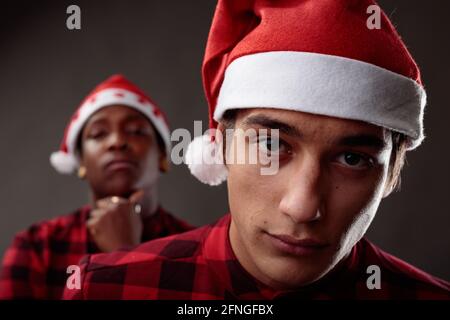 Studio di due giovani che celebrano il Natale indossando rosso Camicie e cappelli di Santa che si pongono uno dietro l'altro con espressione riflessiva simile Foto Stock