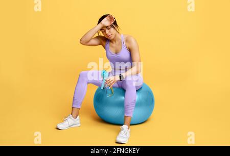 Riposo e relax. Donna nera stanca sportiva seduta su flipper con una bottiglia d'acqua dopo l'esercizio, sfondo giallo Foto Stock