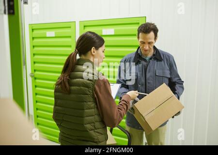 Giovane donna che esamina il documento e le passa la scatola collega mentre lavorano in magazzino Foto Stock