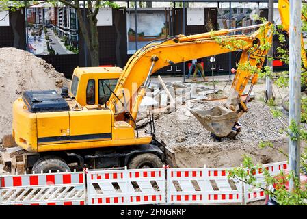 Un escavatore da costruzione pesante lavora su un cantiere recintato e scava una trincea per le utenze urbane. Foto Stock