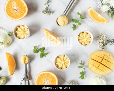 Sorbetto all'arancia e al mango in ciotola e in cucchiai di gelato e spezie di frutta su sfondo marmoreo. Dessert estivo freddo. Vista dall'alto, disposizione piatta Foto Stock