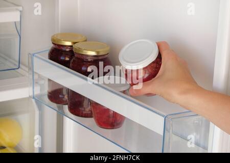 Vasetti di vetro con lampone e mela marmellata su ripiano in frigorifero. Mano femminile con un vaso di marmellata casereccia rossa. Gli alimenti fermentati sani stanno in frigorifero. Foto Stock