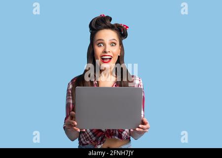 Giovane donna di pinup in abiti retrò che tengono il laptop, sentendosi eccitato sopra la vendita enorme nel webstore, vincendo lotteria in linea Foto Stock