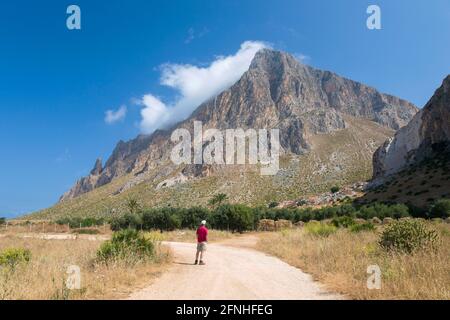 Custonaci, Trapani, Sicilia, Italia. Solitario visitatore su pista polverosa tra i campi ammirando la vista dell'imponente parete sud del Monte cofano. Foto Stock