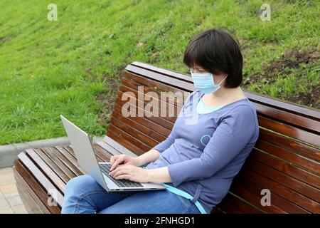 Donna in una maschera medica seduta con un computer portatile in grembo su una panchina di legno nel parco. Concetto di lavoro remoto all'aperto durante un coronavirus COVID-19 Foto Stock