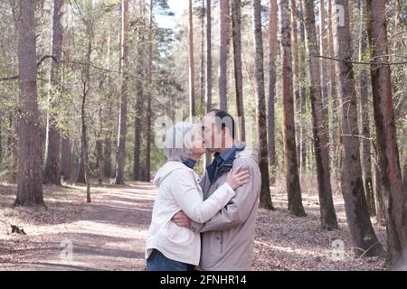 Anziana coppia interracial in abiti casual abbracciandosi l'un l'altro in un parco di foresta primaverile. Una donna che bacia un uomo sulla guancia. Foto Stock