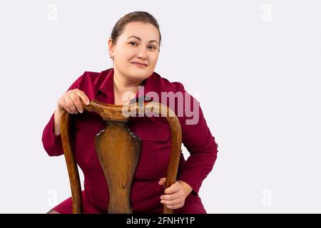 Ritratto di una bella giovane aggraziata più taglia, grande donna su sfondo grigio. La donna di affari che indossa un abito formale si siede su una sedia di legno, in posa dentro Foto Stock