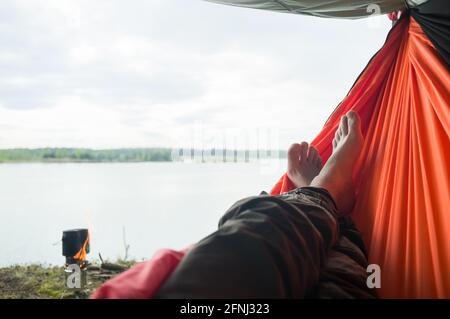 L'uomo spensierato si rilassa sdraiato in un'amaca, sullo sfondo c'è un bruciatore sulla riva di un lago e il cibo viene preparato in una pentola. Foto Stock