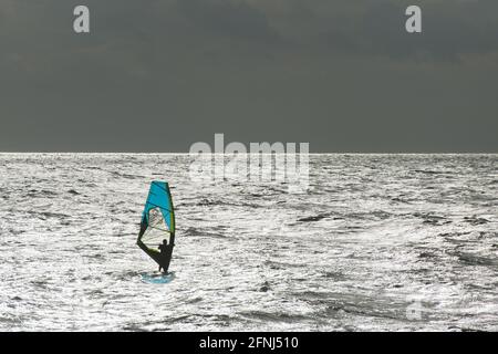 Immagine memorabile di un singolo windsurf con vela blu un mare argentato con orizzonte argentato che separa lo sfondo del cielo scuro Foto Stock