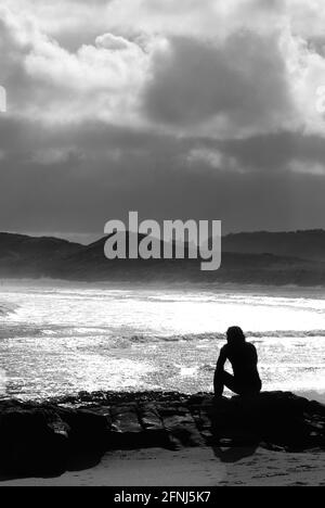 Un'immagine monocromatica in formato verticale di una singola figura in silhouette seduta su rocce in un ambiente costiero con sfondo di dune e nuvole in deriva Foto Stock