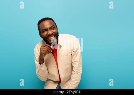 uomo afro-americano malizioso sorridente mentre si guarda lontano isolato sopra blu Foto Stock