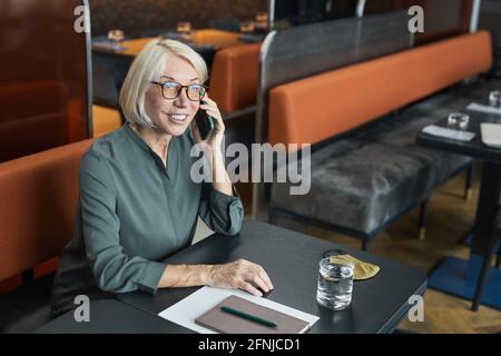 Donna d'affari matura sorridente in occhiali e camicia grigia seduta al tavolo con organizzatore e comunicante per telefono in ristorante Foto Stock