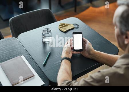 Vista a spalla di un uomo anziano seduto al tavolo con un bicchiere d'acqua e maschera in tessuto e rete da surf sul telefono, schermo vuoto Foto Stock