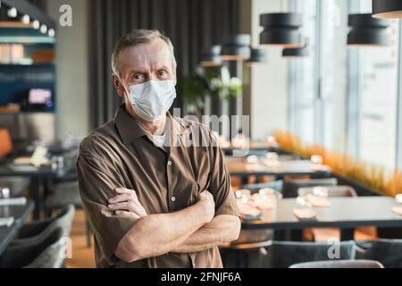 Ritratto di un manager di ristorante fidato in maschera e marrone camicia in piedi con braccia incrociate contro file di tavoli Foto Stock