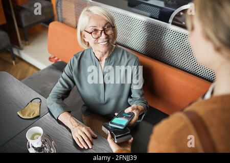Positivo caffè maturo ospite in occhiali seduti al tavolo con maschera in tessuto e a pagamento per il pranzo con gadget in caffetteria Foto Stock