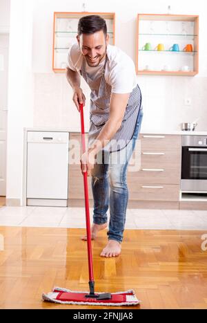 Bel ragazzo che mopping pavimento in parquet di fronte a mobili da cucina e piano di lavoro a casa Foto Stock