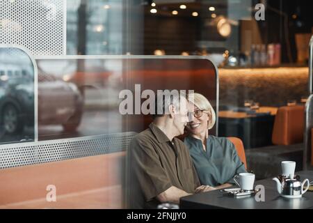 Sorridente affettuosa coppia caucasica senior seduta insieme sul divano e. abbracciarsi mentre si guarda negli occhi l'uno dell'altro in caffetteria Foto Stock