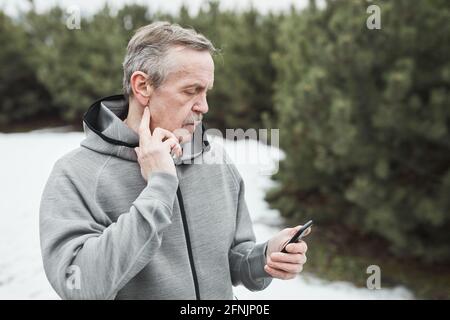 Uomo anziano serio con baffi in piedi contro gli abeti e. controllo del polso tramite cronometro sul telefono Foto Stock
