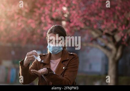 Giovane bella donna con maschera facciale che tiene gel disinfettante e. lavando le mani di fronte all'albero in fiore in primavera Foto Stock