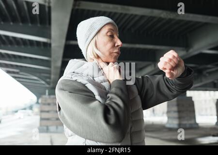 Donna caucasica senior focalizzata con cappello grigio e gilet a contatto collo mentre si prende il polso utilizzando il orologio da polso sotto il ponte all'aperto Foto Stock