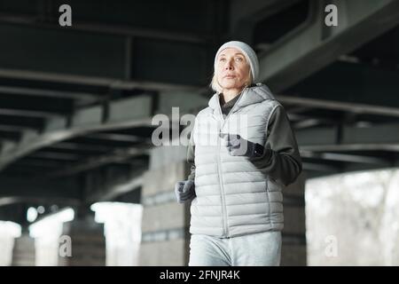Donna matura e sportiva con cappello e gilet da running da solo sotto il ponte mentre si allenano a basse temperature Foto Stock