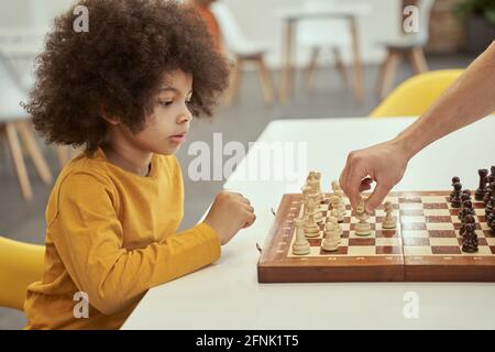 Giocatore entusiasta. Carino ragazzino intelligente pensare mentre si gioca a scacchi con l'adulto, seduto al tavolo all'interno Foto Stock