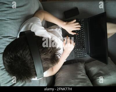 Ragazzo di 15 anni in una T-shirt bianca si siede sopra il divano con un computer portatile Foto Stock