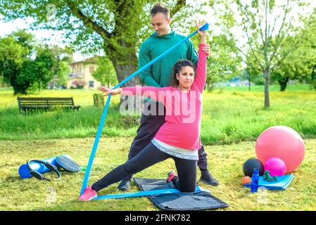 Giovane donna incinta allenamento braccia con elastico resistenza corda elasticizzata seduta su una palla da palestra. Personal trainer allenare una ragazza all'aperto nel parco. Foto Stock