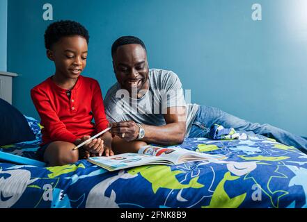 Il padre sorridente aiuta il figlio sorridente con i compiti sul letto a casa Foto Stock