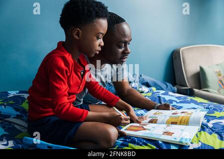 Padre che aiuta il figlio con i compiti sul letto a casa Foto Stock