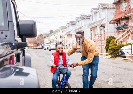 Il padre sorridente che aiuta il figlio a guidare una bicicletta sul strada fuori Foto Stock