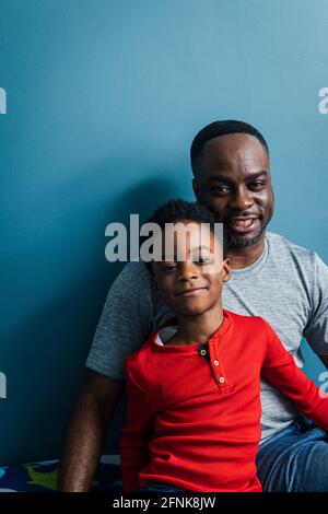Ritratto di padre sorridente e figlio seduto contro la parete blu a casa Foto Stock