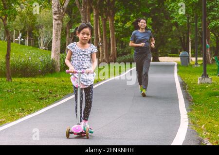 Bella infanzia, insieme con i genitori nel parco, l'esercizio con la madre, bambina asiatica in sella scooter e madre jogging seguire sulla strada nel parco Foto Stock