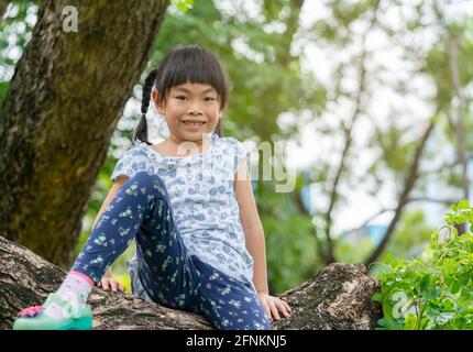 La bambina asiatica è seduta sul brunch del grande albero, guardando la macchina fotografica, lo spazio per il testo. Foto Stock