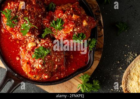 Vista ravvicinata di tre polpette tradizionali italiane di grandi dimensioni in salsa di pomodoro sfrigolante in padella di ghisa. La padella si siede su trivet di legno e foglie di prezzemolo Foto Stock