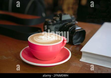 Tazza di caffè in tazzina rossa e libro, con macchina fotografica sullo sfondo su tavola di legno. Foto Stock