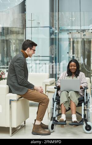 Donna africana disabile seduta in sedia a rotelle e che lavora su un computer portatile con il suo collega seduto accanto a lei in ufficio Foto Stock