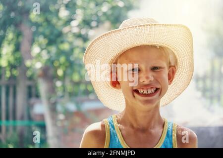 Ragazzo allegro che indossa un grande cappello di paglia e senza maniche blu una camicia che si posa per la macchina fotografica e sorride in bocca nel soleggiato parco cittadino estivo vicino Foto Stock