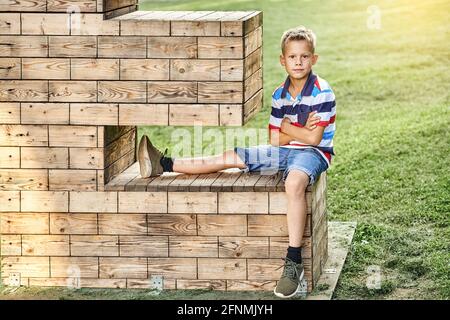 Bambino positivo in polo a righe t-shirt e jeans shorts siede su installazione decorativa in legno su un lussureggiante prato verde parcheggio Foto Stock
